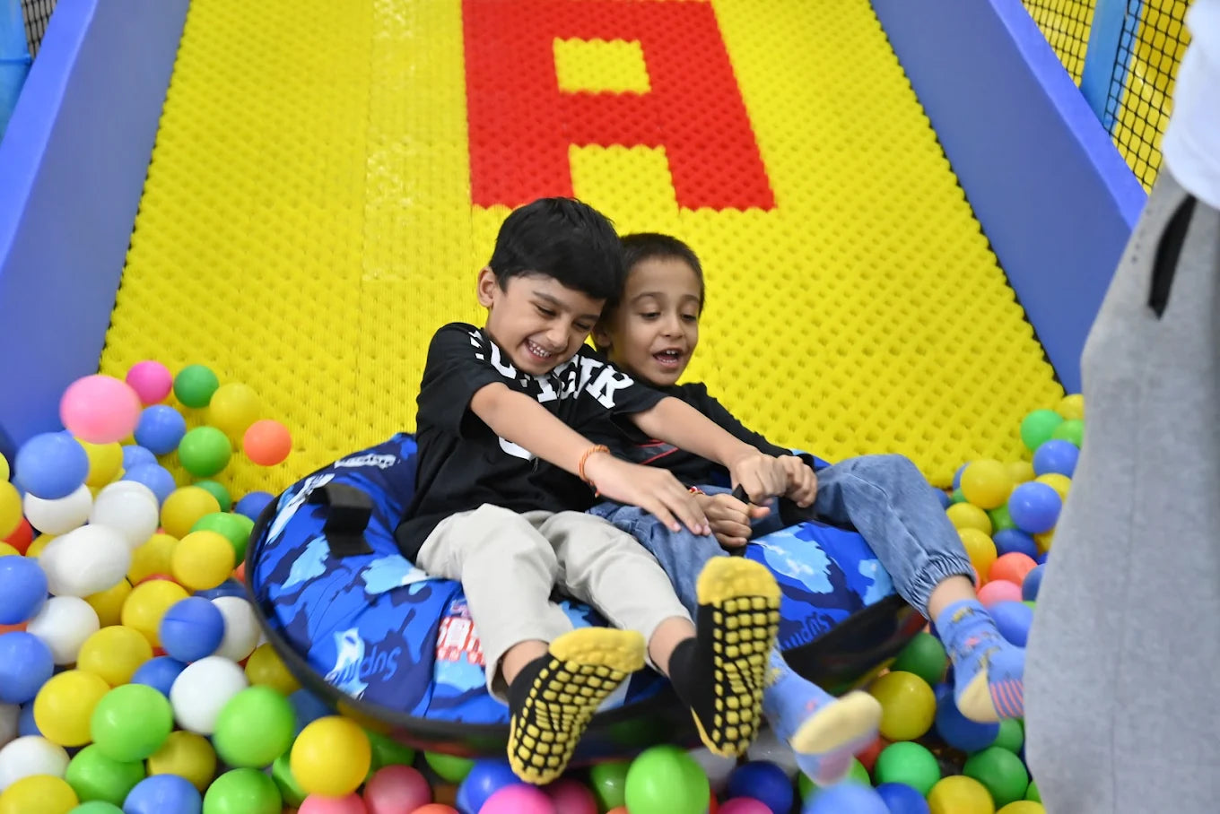 Two young boys slide down a colorful ball pit on a tube.