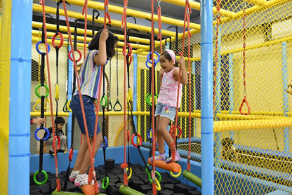 Two young girls navigate a colorful indoor obstacle course with ropes and rings.