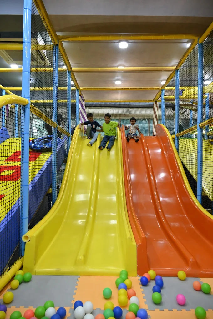 Three young boys slide down a yellow and orange playground slide.