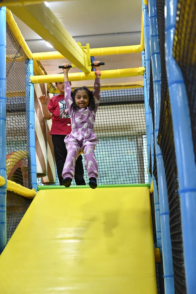 A young girl swings on monkey bars in a colorful indoor playground, with a boy behind her.