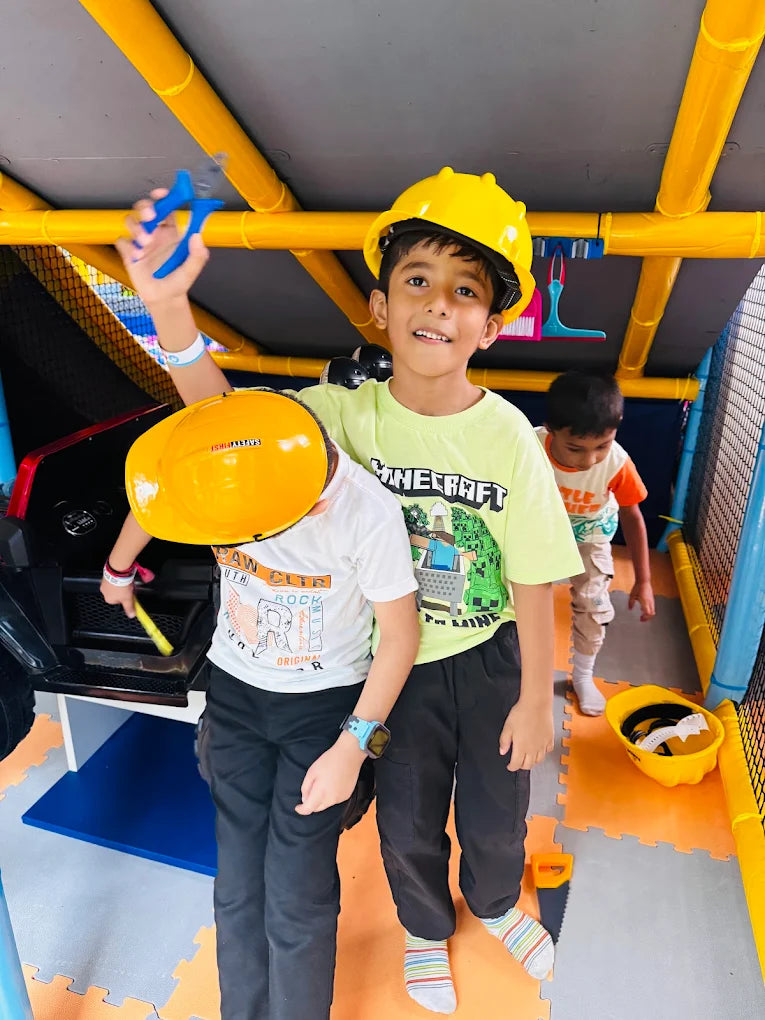 Two young boys in hard hats play with toy tools in a colorful indoor play area.
