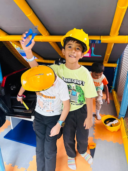 Two young boys in hard hats play with toy tools in a colorful indoor play area.