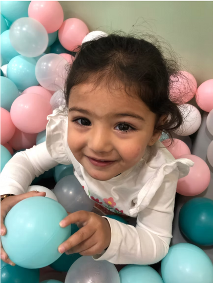 A young girl smiles while playing in a ball pit filled with pink, blue, and white balls.