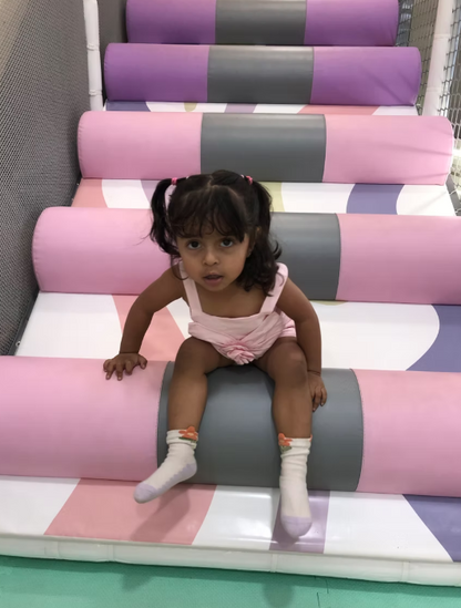 A young girl sits on a padded play structure with colorful cylindrical steps. She is wearing a pink outfit and white socks.