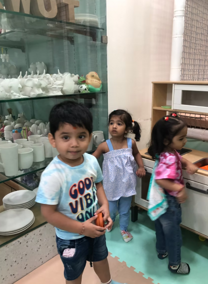Three young children stand in a room with shelves of white ceramic figures and a toy kitchen.