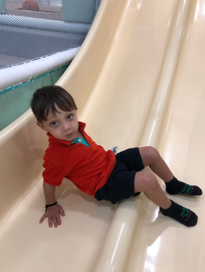 A young boy sits on a cream-colored slide, wearing an orange shirt and dark shorts.