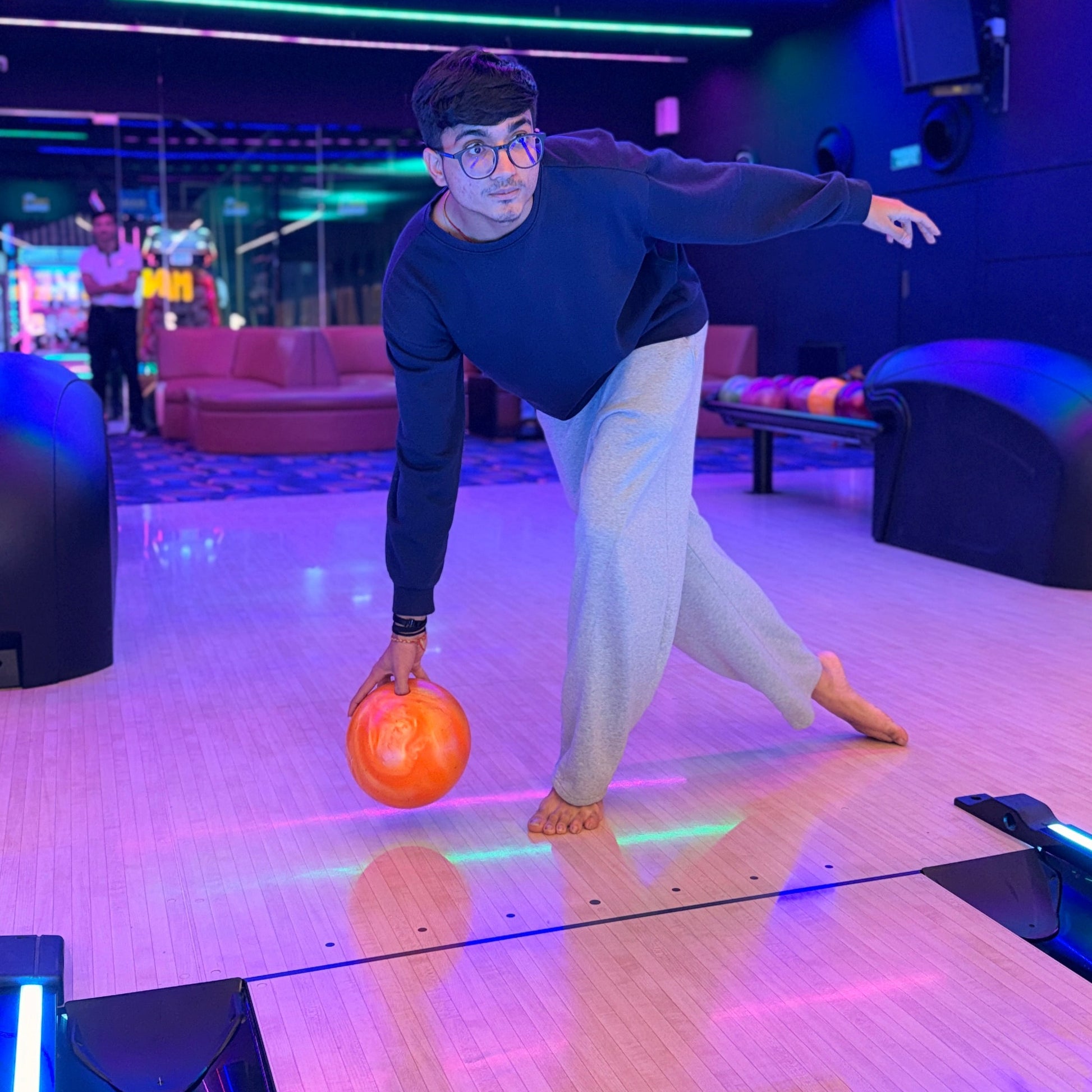A young man in a bowling alley prepares to bowl an orange ball. The lane is lit with colorful neon lights.