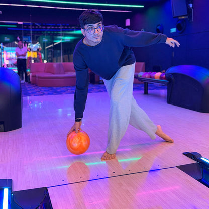 A young man in a bowling alley prepares to bowl an orange ball. The lane is lit with colorful neon lights.