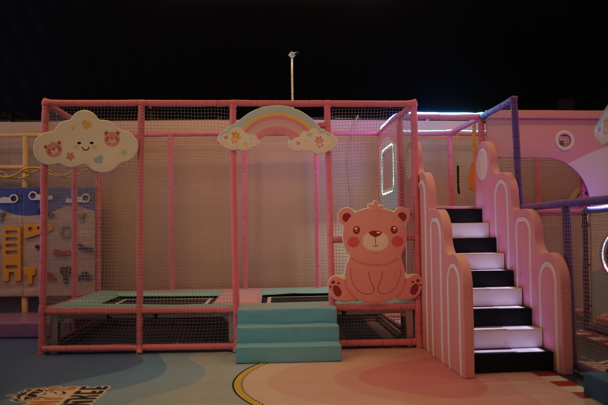 A brightly colored indoor playground with a trampoline, slide, and climbing wall for children.