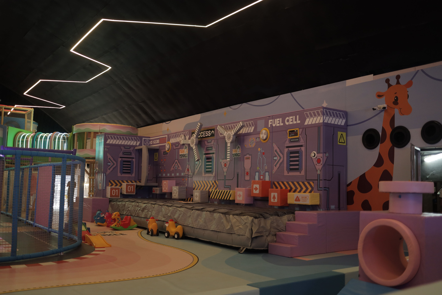 A colorful indoor playground with a giraffe mural and a "Fuel Cell" themed play structure.