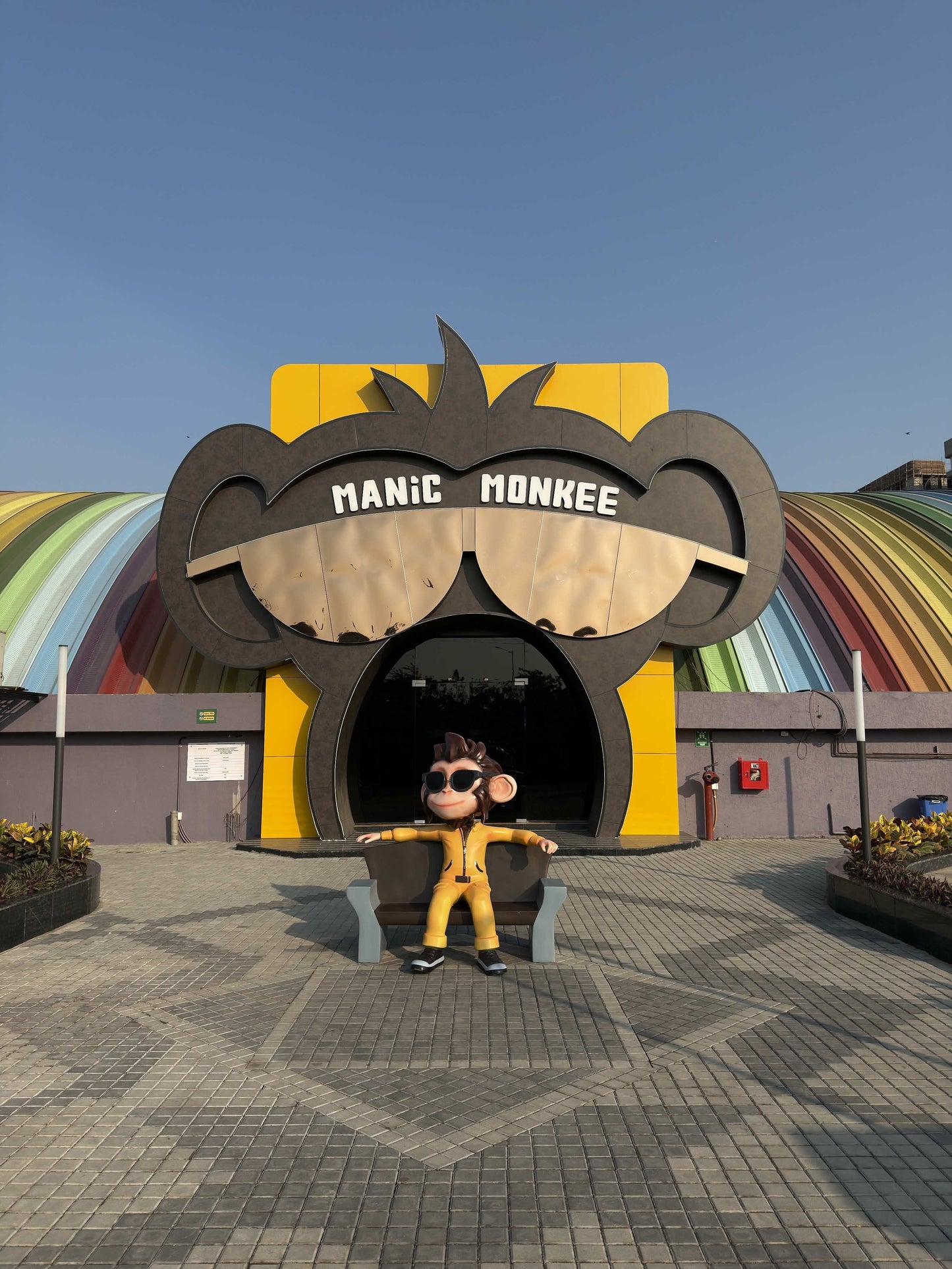 A statue of a monkey in a yellow jumpsuit sits on a bench in front of a building shaped like a monkey's face.