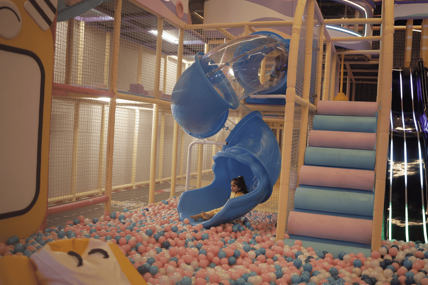 A young child slides down a blue tube into a ball pit in a colorful indoor playground.