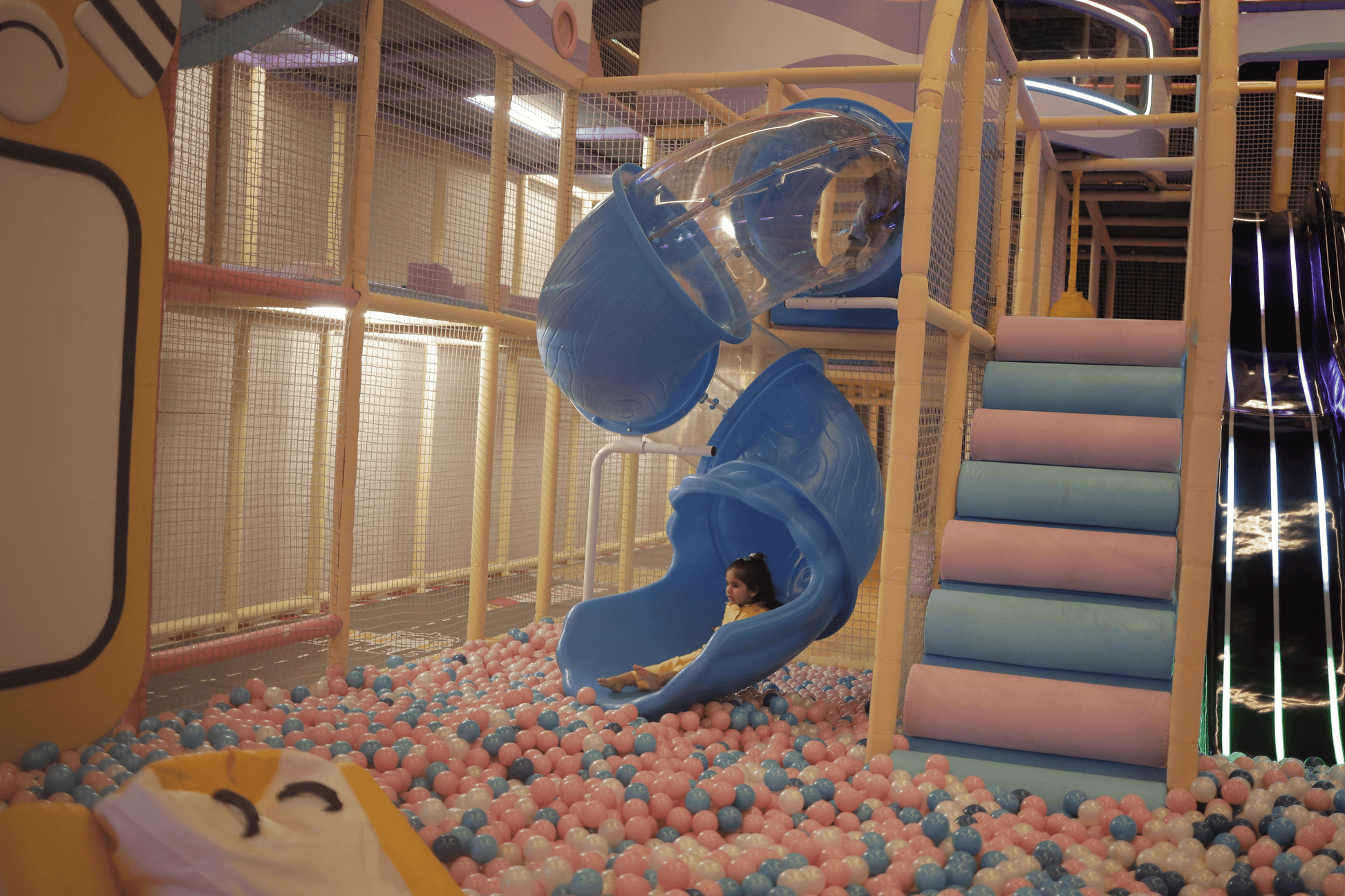 A young child slides down a blue tube into a ball pit in a colorful indoor playground.