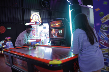 Two young adults play an air hockey game in a brightly lit arcade.