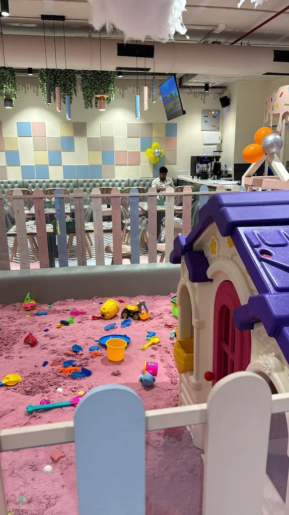 A child plays in a pink sandpit filled with toys, with a playhouse and seating area in the background.