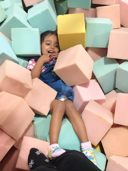 A young child smiles while sitting in a ball pit filled with colorful foam blocks.