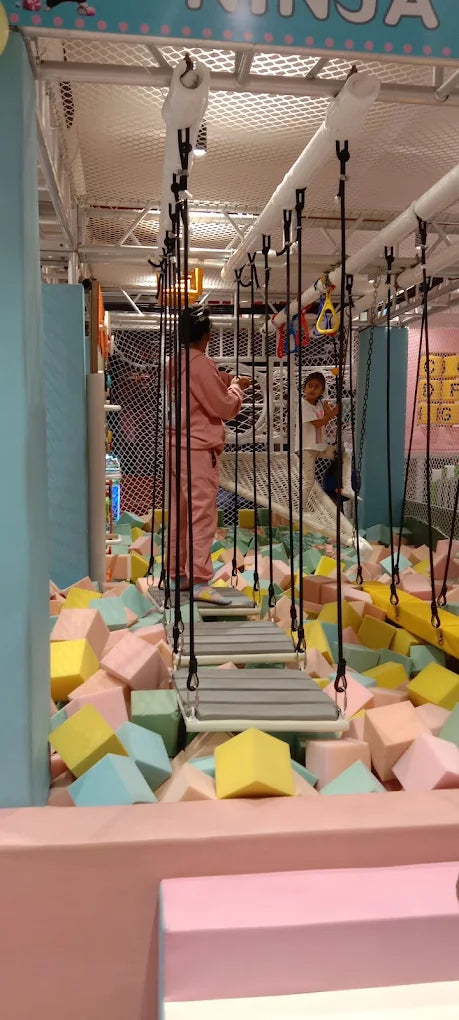 A child navigates a rope bridge over a foam pit at an indoor playground.