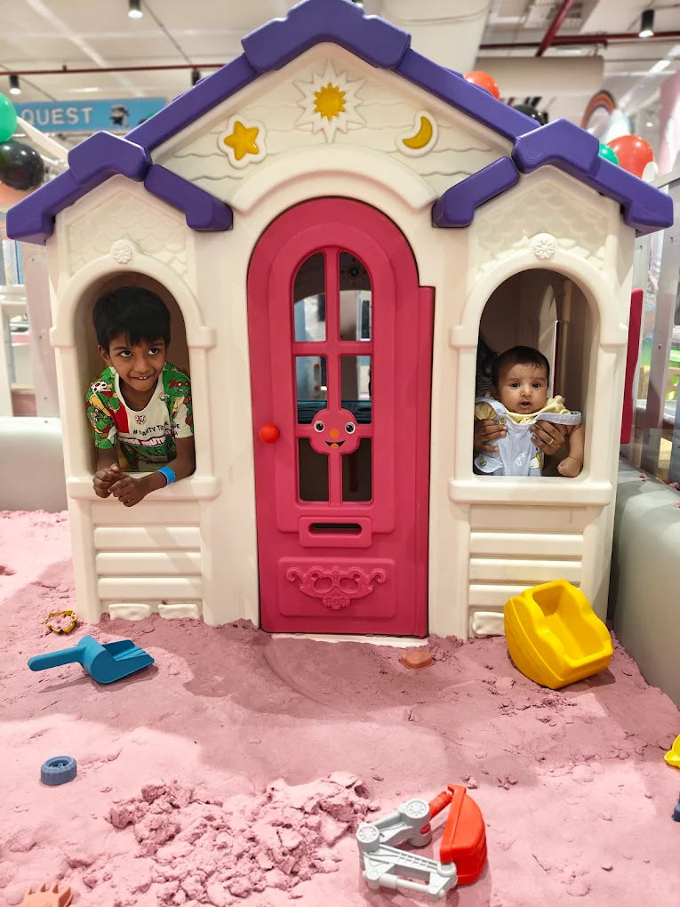 Two young children play in a pink sandbox in front of a toy house.