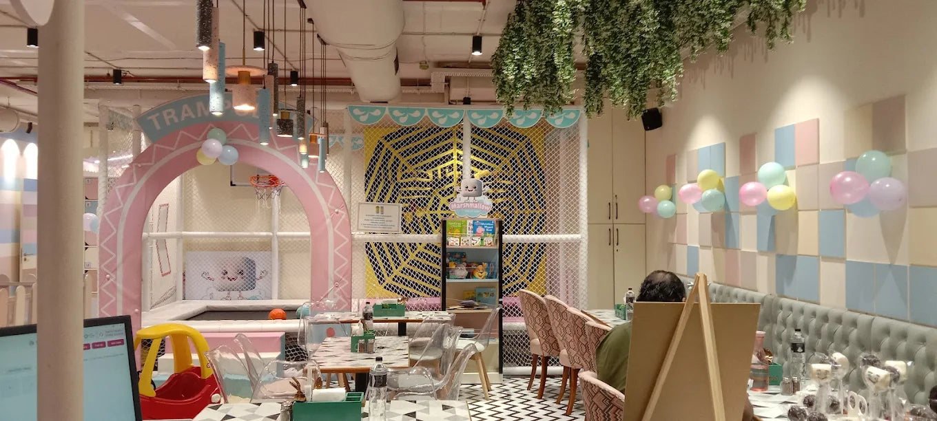 A brightly decorated indoor play area for children with tables and chairs. A person is seated at a table in the foreground.