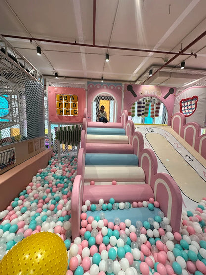 A child plays in a ball pit next to a pink and blue slide in an indoor playground.