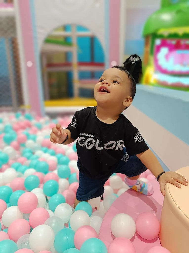 A young child smiles while playing in a ball pit. The child is wearing a black t-shirt and blue shorts.