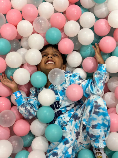 A young child smiles while lying in a ball pit filled with pink, white, and blue balls.