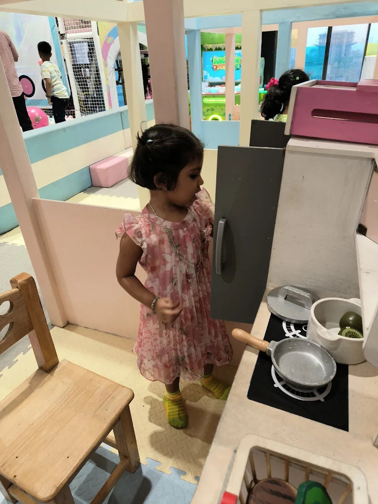 A young girl plays in a toy kitchen at an indoor playground. She wears a pink dress and yellow socks.