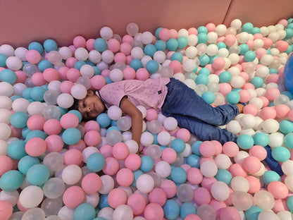 A young child lies in a ball pit filled with pink, blue, and white balls.