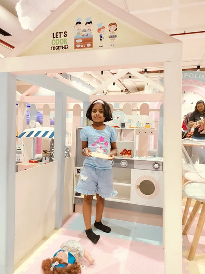 A young girl stands in a play kitchen holding a plate, with a doll on the floor.