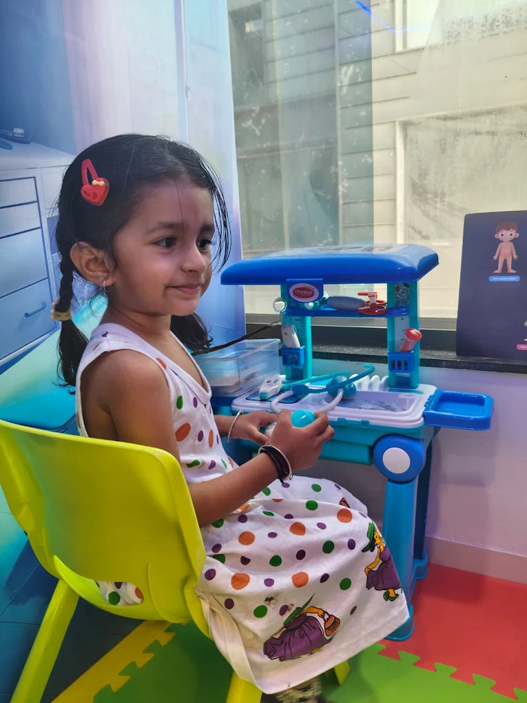 A young girl sits at a toy doctor's desk, playing with medical tools.