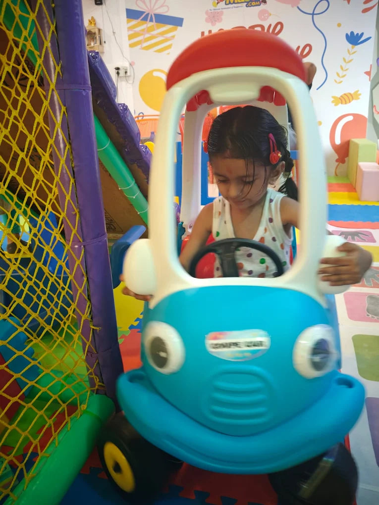 A young girl plays in a toy car inside a colorful indoor playground.