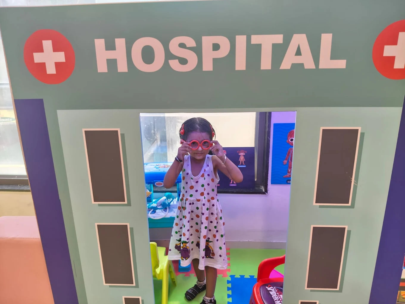 A young girl plays dress-up in a toy hospital, wearing heart-shaped glasses.