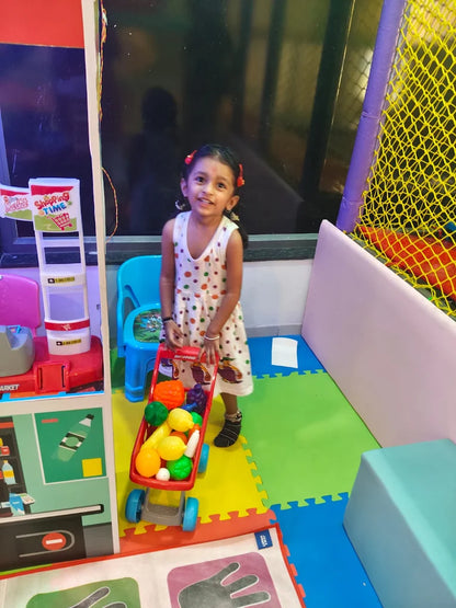 A young girl plays with a toy shopping cart filled with plastic fruit in a colorful indoor play area.