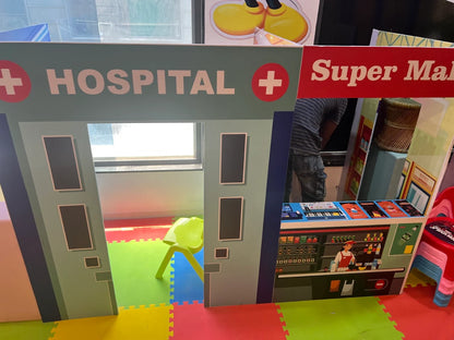 A child's play area with a hospital and supermarket facade, featuring colorful mats and a plastic chair.