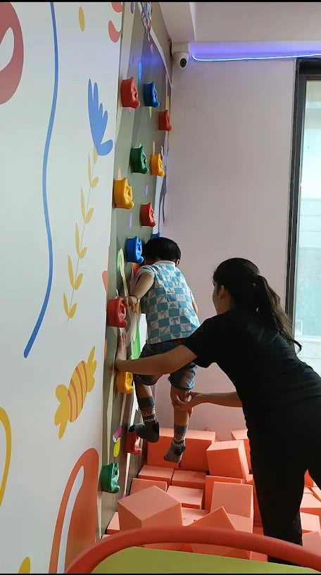A child climbs a colorful wall with an adult spotting them, surrounded by soft play blocks.