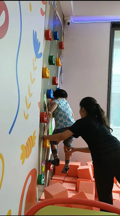 A child climbs a colorful wall with an adult spotting them, surrounded by soft play blocks.