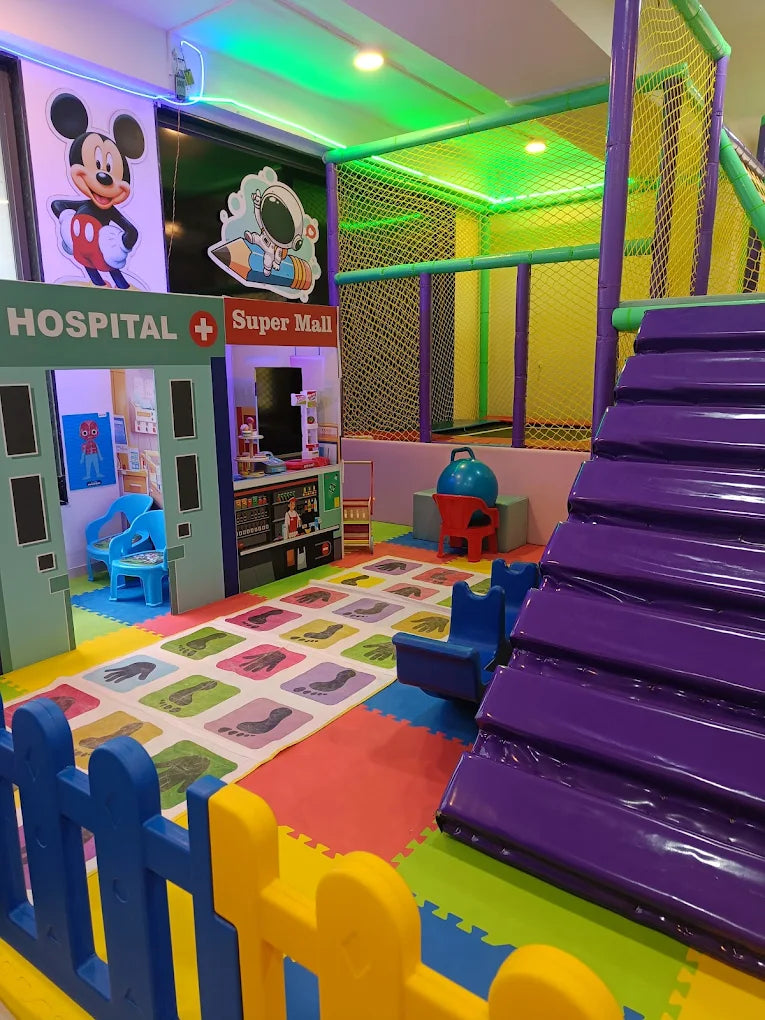 A colorful indoor playground with a hospital and mall theme, featuring a soft play structure and a mat with foot imprints.
