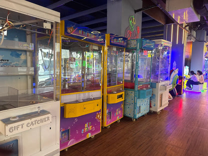 A row of colorful claw machines in an arcade, with young adults playing in the background.