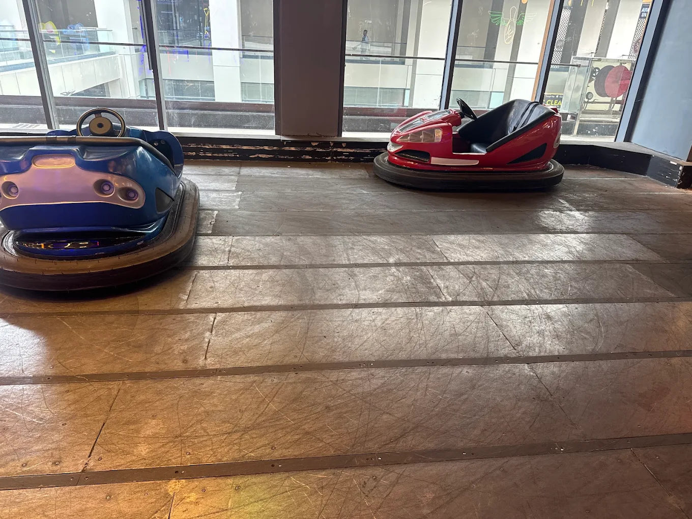 Two bumper cars, one blue and one red, sit on a wooden floor in an indoor amusement area.