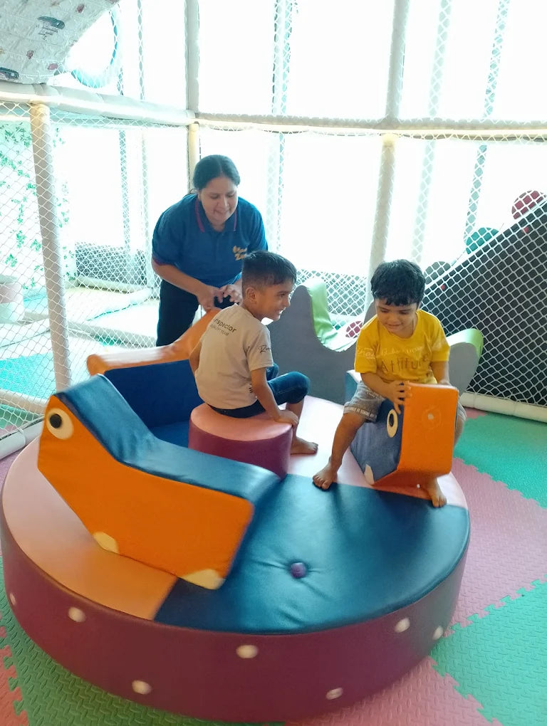Children playing on colorful toy cars in a playground with an adult supervising.