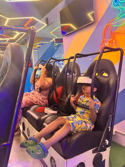 Two children wearing VR headsets sit in a virtual reality ride. The ride has a futuristic design with neon lights.