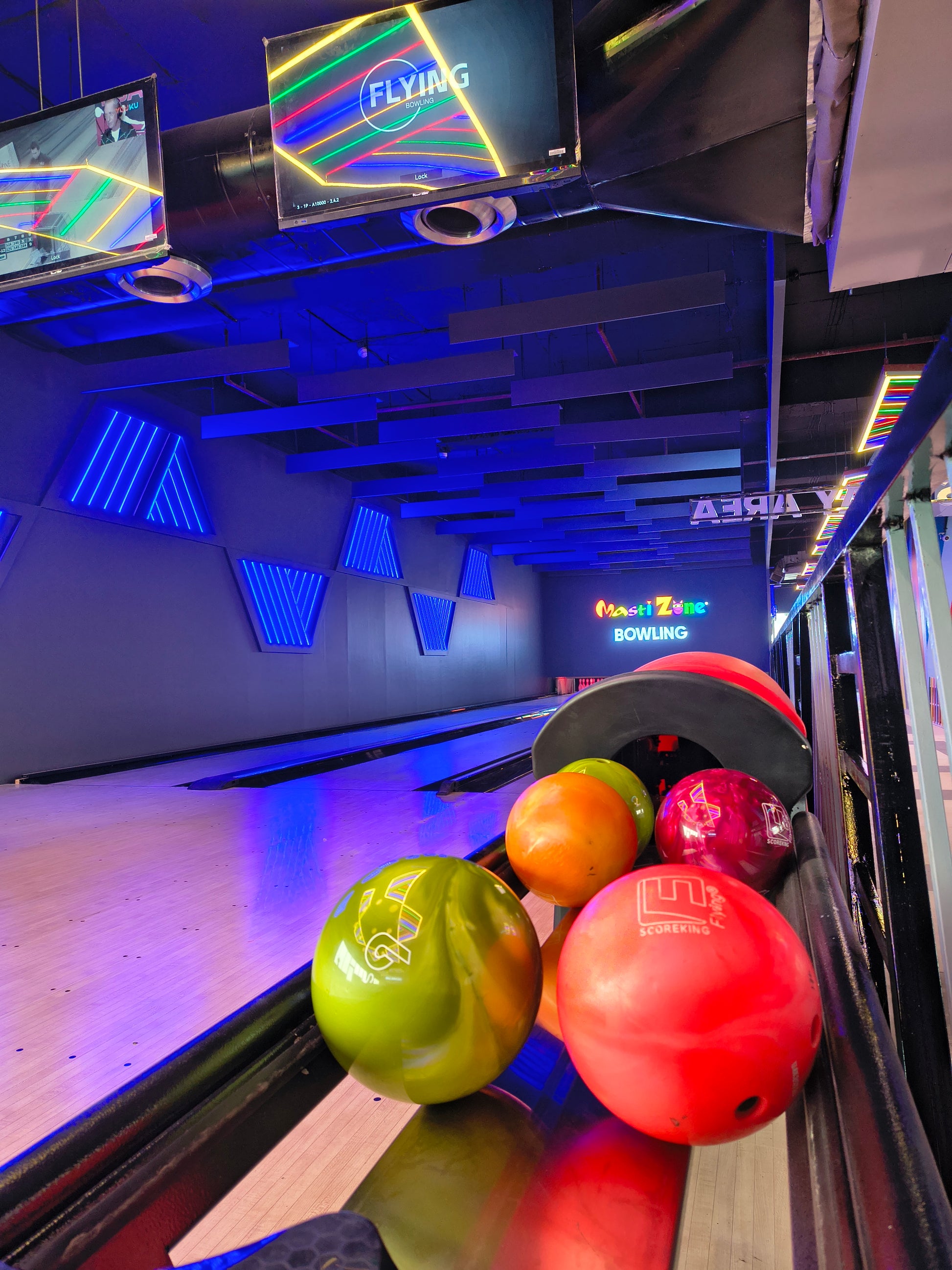Bowling balls in various colors sit on a rack at a bowling alley with neon lights.
