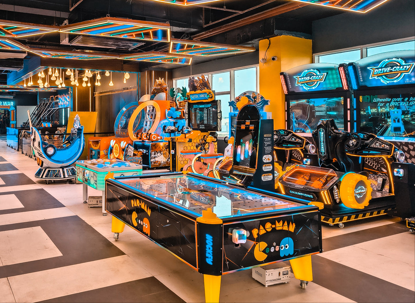 An arcade with various games, including racing simulators and a Pac-Man hockey table, illuminated by neon lights.