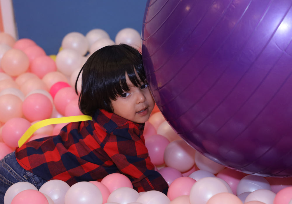 A young child peeks out from behind a large purple ball in a ball pit filled with pink and white balloons.