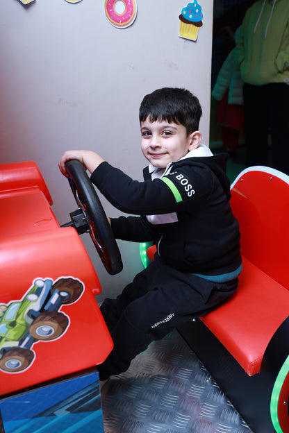 A young boy smiles while pretending to drive a red toy car. He is wearing a black hoodie and pants.