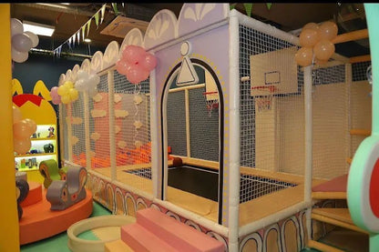 A colorful indoor playground with soft play structures, balloons, and a basketball hoop. Designed for young children's active play.