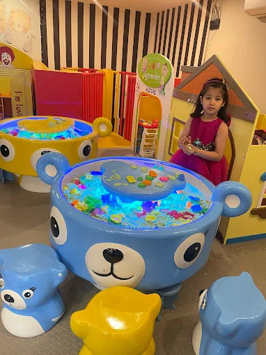 A young girl in a pink dress plays in a colorful indoor playground with a blue bear-shaped water table.