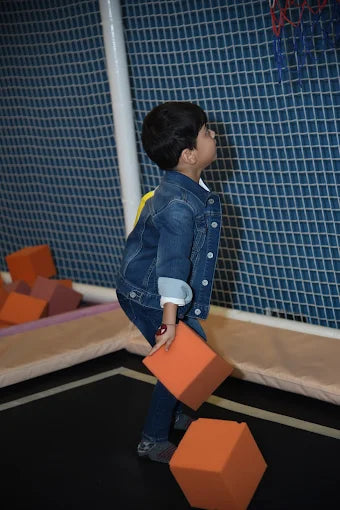 A young boy in a denim jacket holds an orange cube in a play area with soft blocks and a net wall.