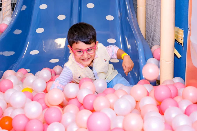 A young boy in pink glasses plays in a ball pit with a blue slide.