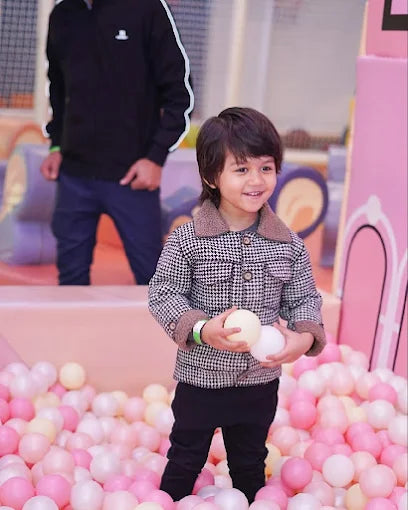 A young boy smiles while holding balls in a ball pit, with an adult in the background.
